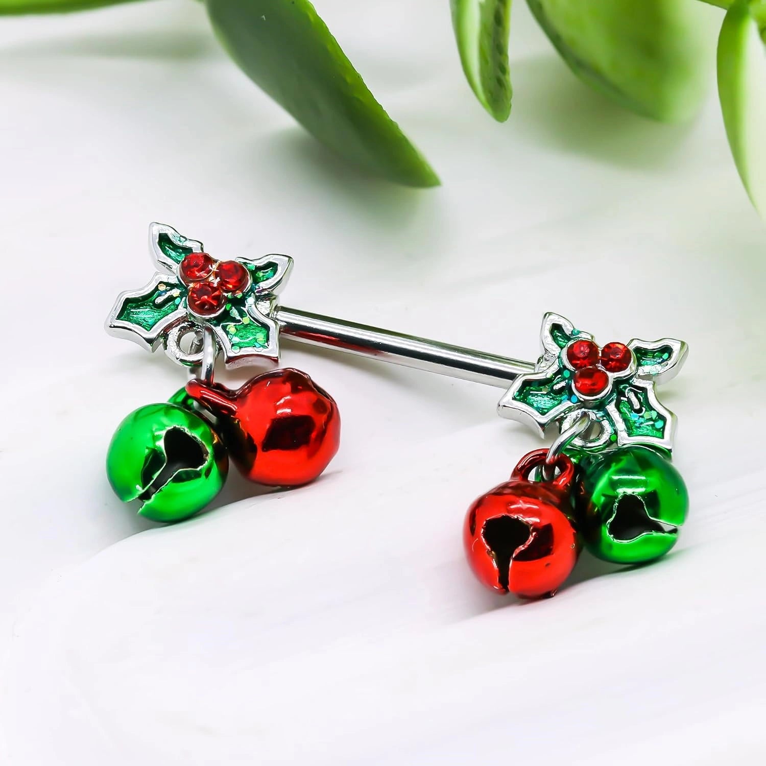 A pair of shiny stainless steel nipple barbells with dangling Christmas bells, displayed on a festive red background.