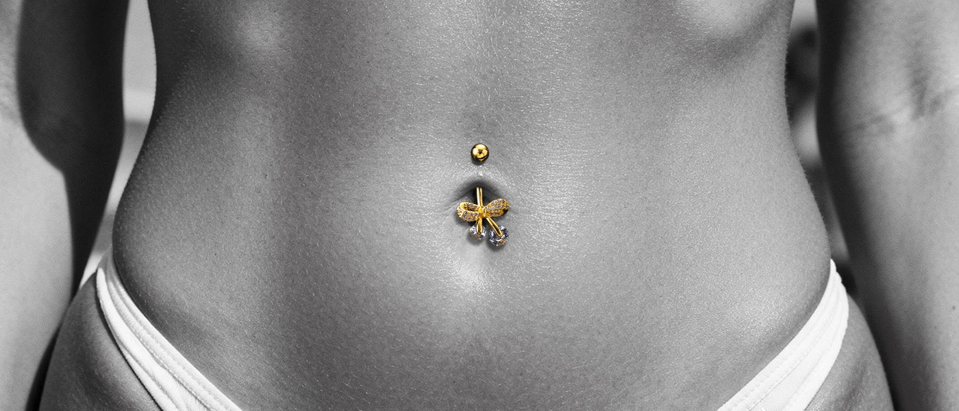 A close-up photo showcasing the gold belly ring on a model. The fixed, diamond-encrusted bow design is clearly visible on the lower end.